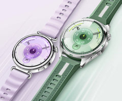 Two smartwatches with round silver-toned cases shown on a pale mint background. One watch has a light lavender strap and a purple dial, and the other has a green strap and a green dial. huawei watch gt 6